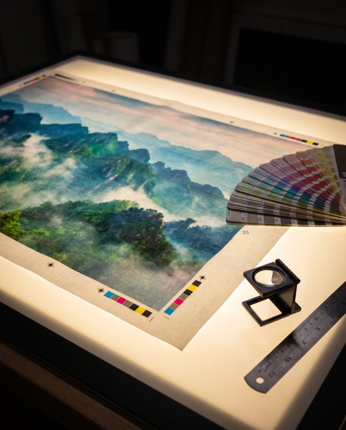 Large-format fabric print proof of a mountain graphic on a light table with colour bars, swatches and a loupe